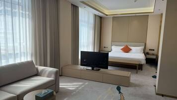 A modern hotel room with a sofa, a television on a stand, a bed with an orange pillow, and a large window with sheer curtains.