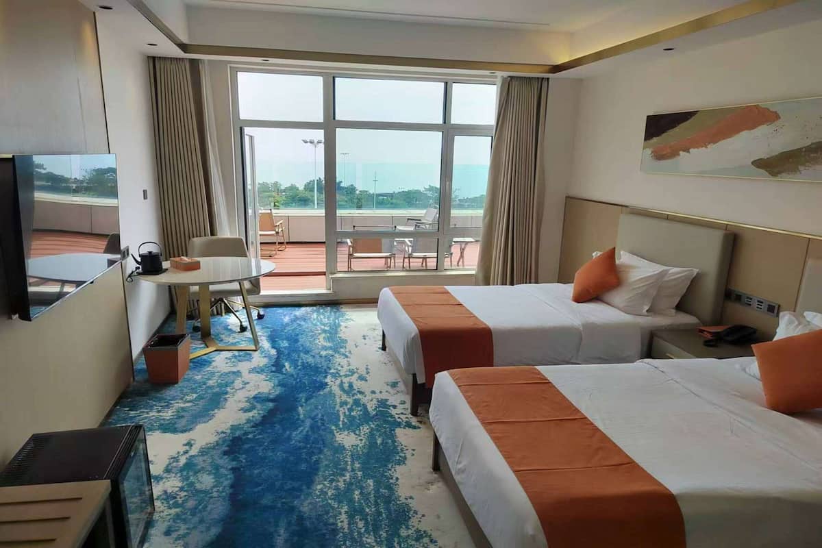A hotel room with two beds, a desk, a chair, and a large window offering a view of the outdoors.