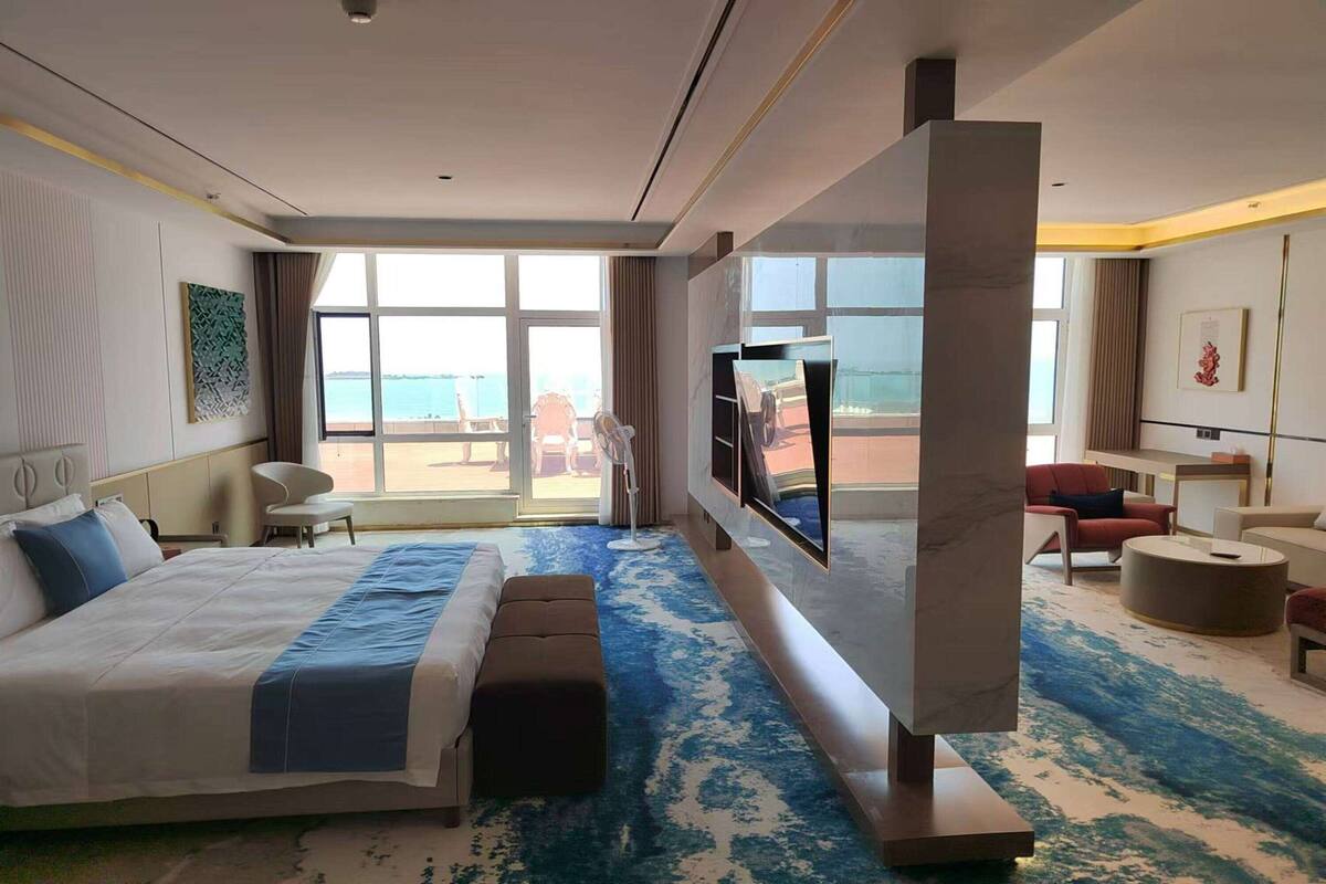 A hotel room with a large bed, a flat-screen TV, a seating area with a chair and ottoman, and a view of the sea through large windows.