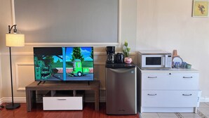 Ett vardagsrum med en platt-TV på ett trästativ, en kaffebryggare, en kaktus, en mikrovågsugn och ett vitt skåp med lådor.