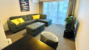 A living room with a sectional sofa, ottoman, and a white chair. There is a plant in a black pot and a painting on the wall. - OK Holiday Unit #5 - Queenstown Holiday Apartment (Queenstown)