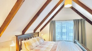 A bedroom with a sloped ceiling, a bed with a wooden headboard, a bedside table with a lamp, and a window with curtains. - OK Holiday Unit #5 - Queenstown Holiday Apartment (Queenstown)