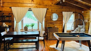 Un intérieur de chalet chaleureux avec un plafond en bois, une table de billard, une table à manger avec des chaises et un fauteuil suspendu.