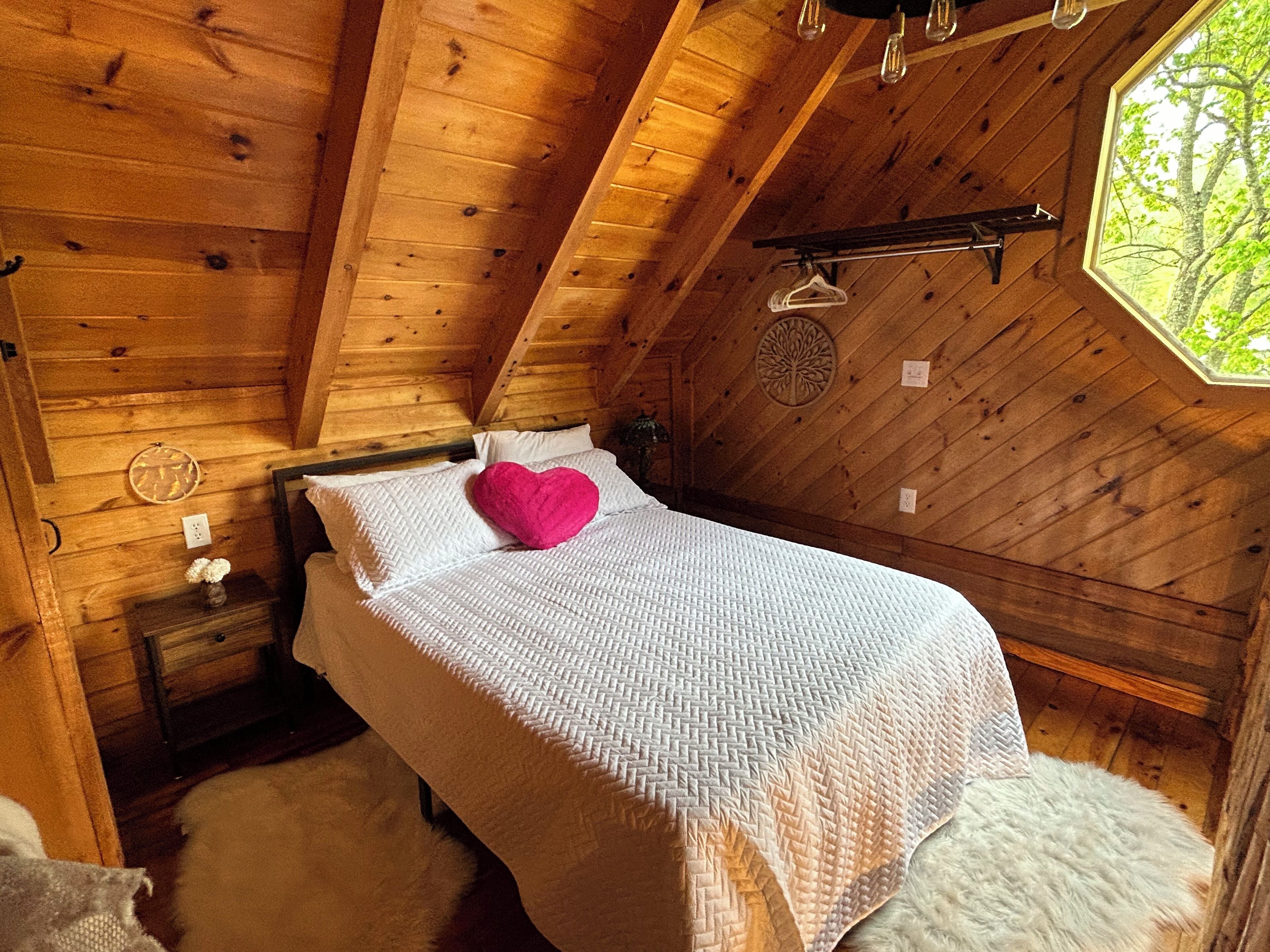 A cozy wooden cabin bedroom with a bed, a hanging clothes rack, and a window with a view of trees.