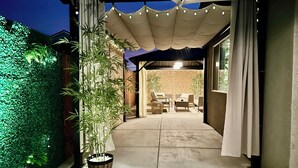 Ein Terrassenbereich im Freien mit Betonboden, einem Kübel mit Bambuspflanze, Korbmöbeln und einer Pergola mit Lichterketten.