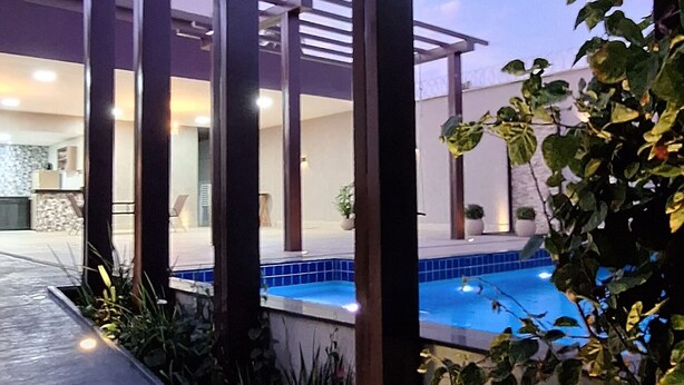 A modern outdoor pool area with a clear blue pool, surrounded by plants and a wooden pergola. - Espaço de Festas e Descanso Cintia Lima (Petrovale Betim)