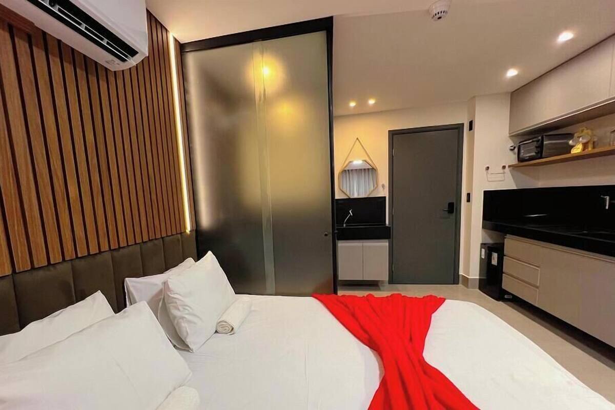 A modern hotel room with a large bed, a wooden panel wall, a sliding door, a kitchenette, and a ceiling-mounted air conditioner.