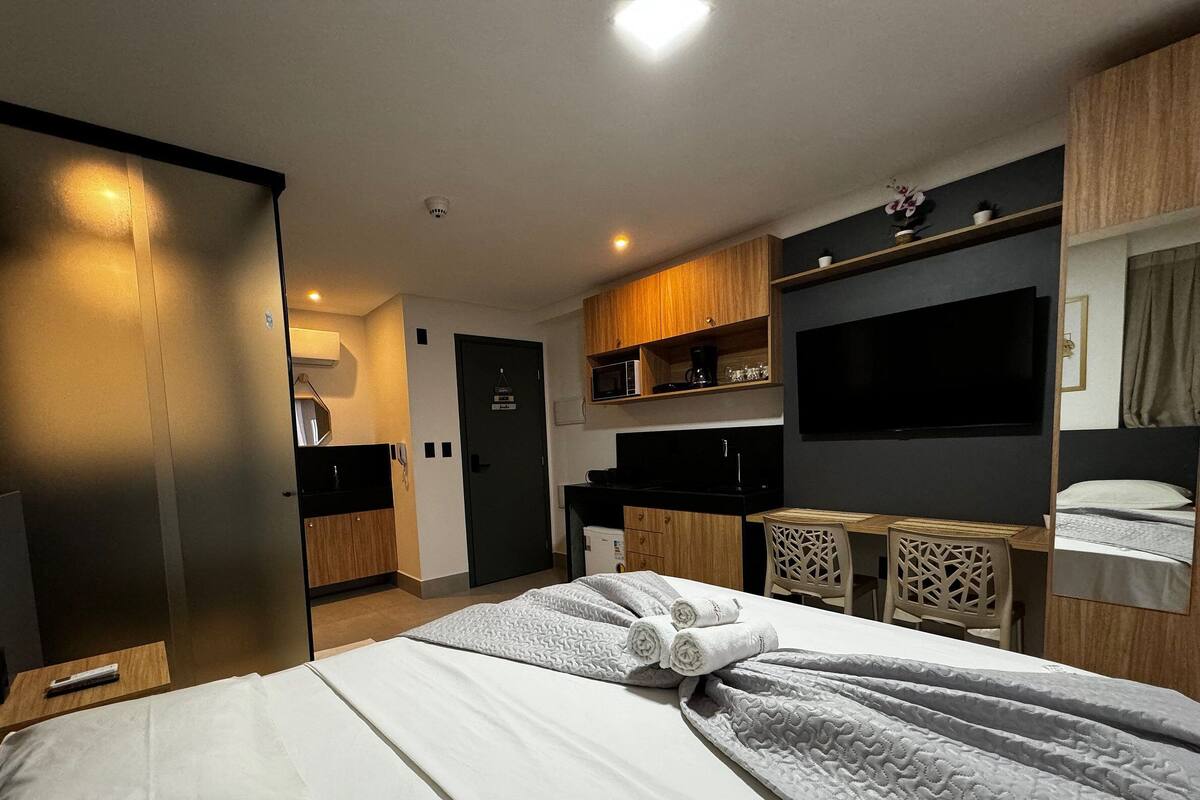 A modern hotel room with a bed, a kitchenette, a TV, and a bathroom.
