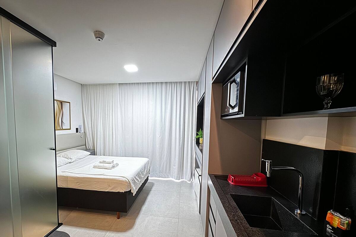 A compact hotel room with a bed, a small bedside table, a mirror, a window with curtains, a kitchenette with a microwave, and a red object on the counter.