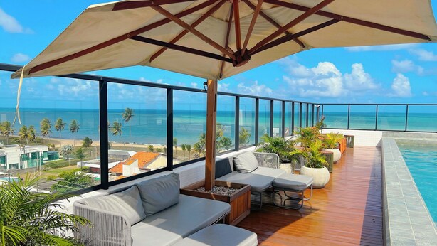 A balcony with a pool, seating, and a view of the ocean. - Get By Side  (João Pessoa)