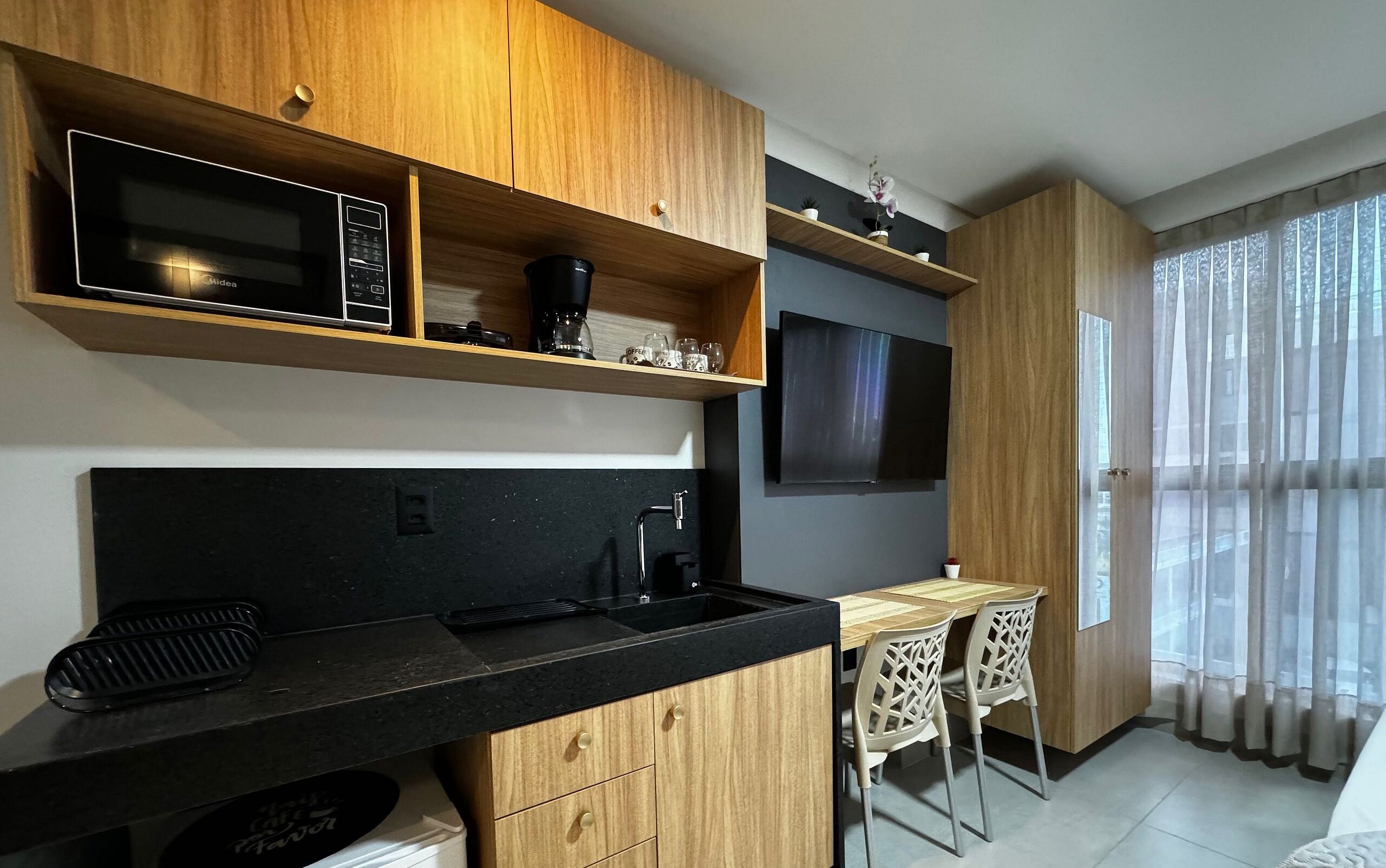 A modern kitchen with wooden cabinets, a black countertop, a microwave, a coffee machine, and a TV mounted on the wall.