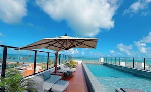 Une piscine sur le toit offrant une vue dégagée sur l’océan, un espace salon couvert avec des coussins et un grand parasol pour faire de l’ombre.