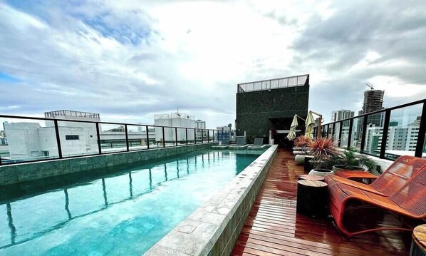Une piscine sur le toit, avec une terrasse en bois, des chaises longues et une vue sur le paysage urbain.