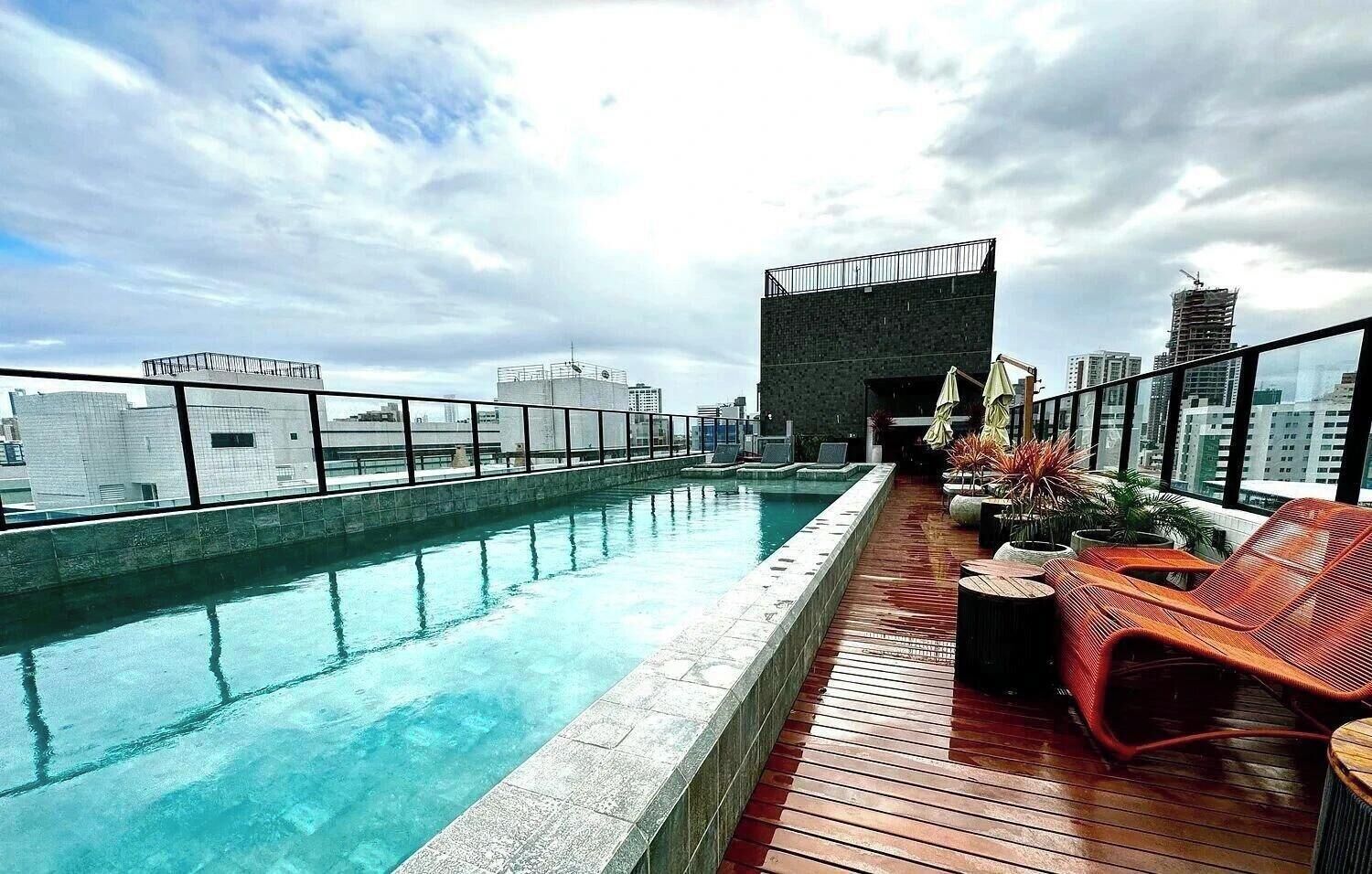 A rooftop pool with a wooden deck, lounge chairs, and a view of the city skyline.