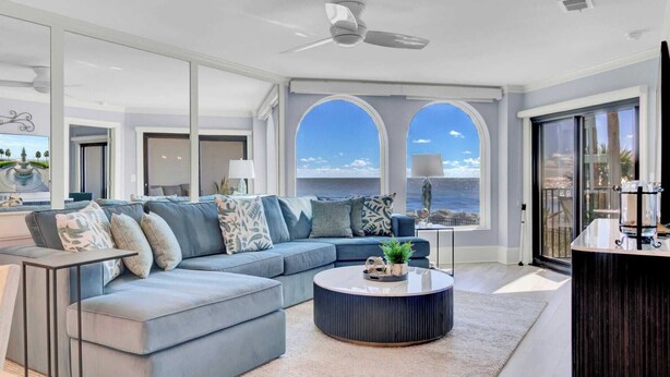 A modern living room with a large blue sofa, a round coffee table, and a view of the ocean through arched windows.