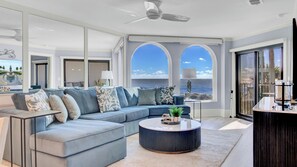 A modern living room with a large sofa, a round coffee table, and a view of the ocean through arched windows. - 118 Seascape by Avantstay Pool + Oceanfront Views (Isle of Palms)