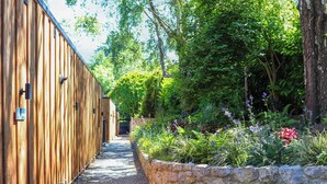 Ein schmaler Pfad, gesäumt von Holzhäusern und einer Steinmauer, mit üppigem Grün und Blumen.