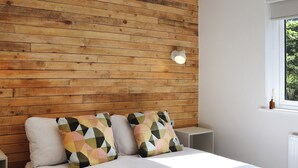 Ein Schlafzimmer mit Holzwand, einem Bett mit gemusterten Kissen, einem Nachttisch und einem Fenster mit Blick ins Grüne.