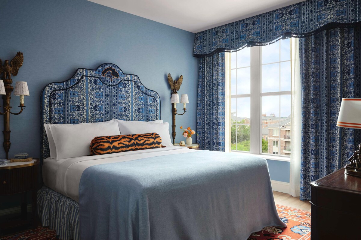A bedroom with a large bed, a window with curtains, a nightstand with a lamp, and a wall-mounted lamp.