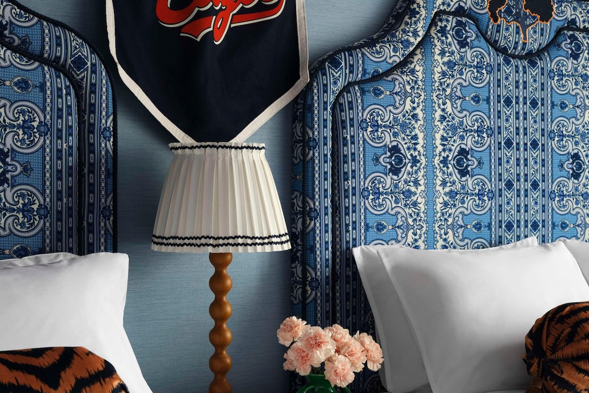 Una camera d'albergo con un letto, un comodino con una lampada, un vaso con fiori e un quadro con la scritta 'Vogue Eagle'.