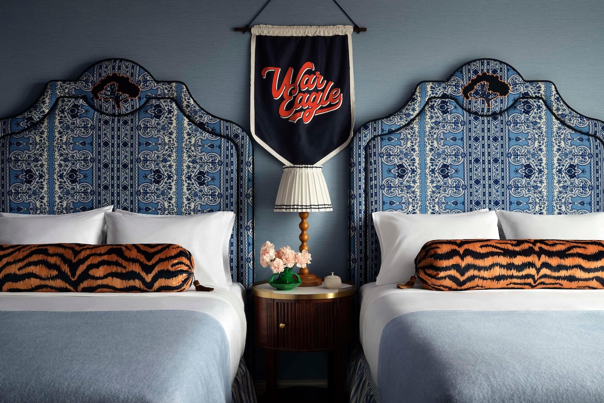 A hotel room with two beds, each with a tiger-striped pillow, a nightstand with a lamp and flowers, and a wall hanging with "War Eagle" text.