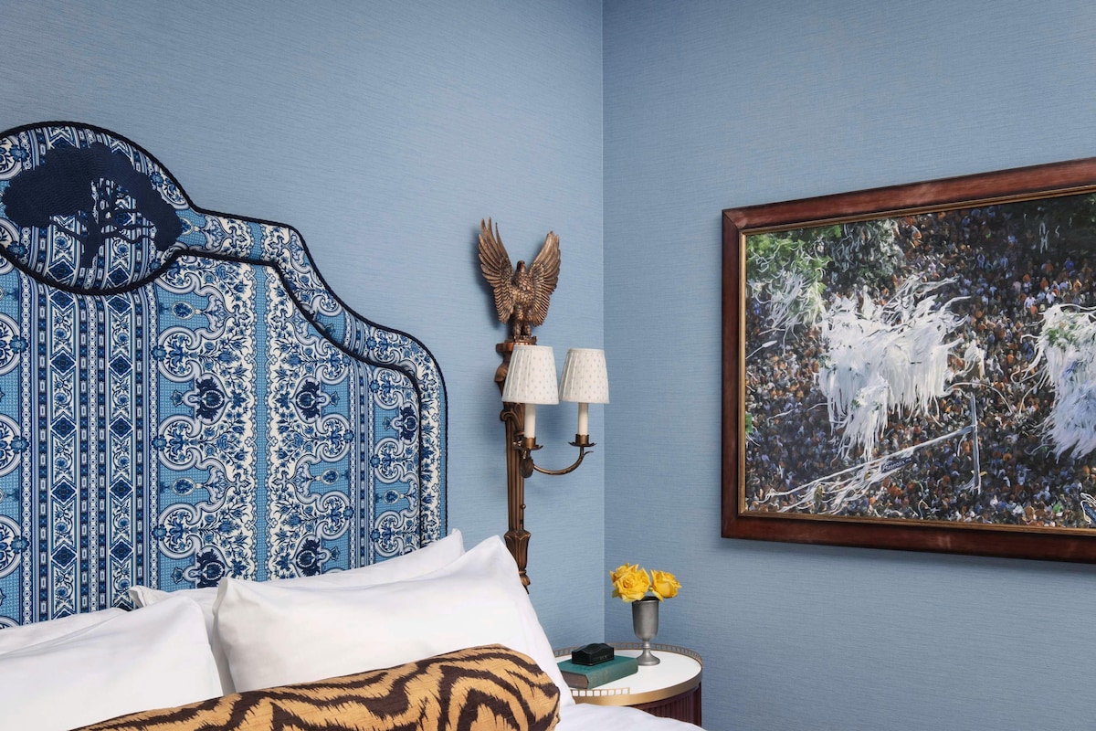 A hotel room with a patterned headboard, a bed with white and brown bedding, a nightstand with a lamp and a vase of yellow flowers, and a framed painting on the wall.