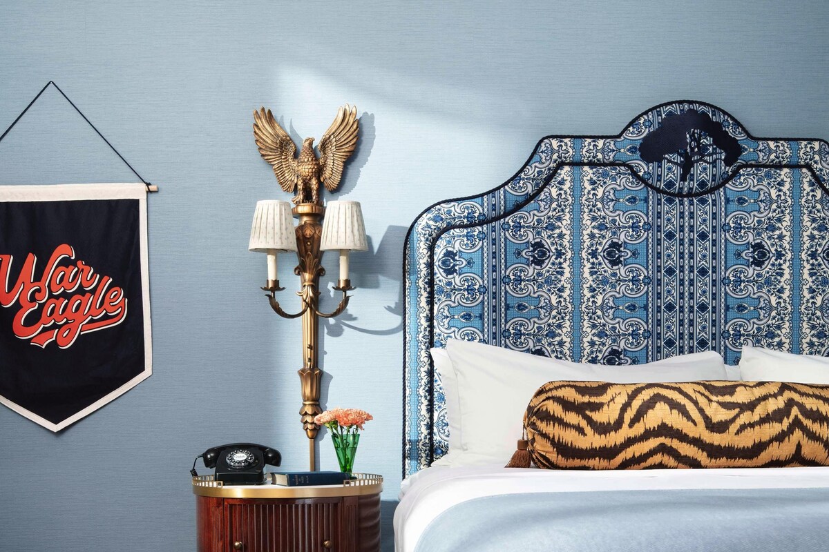 A bedroom with a patterned headboard, a bedside table with a lamp, a framed sign, and a decorative wall piece.