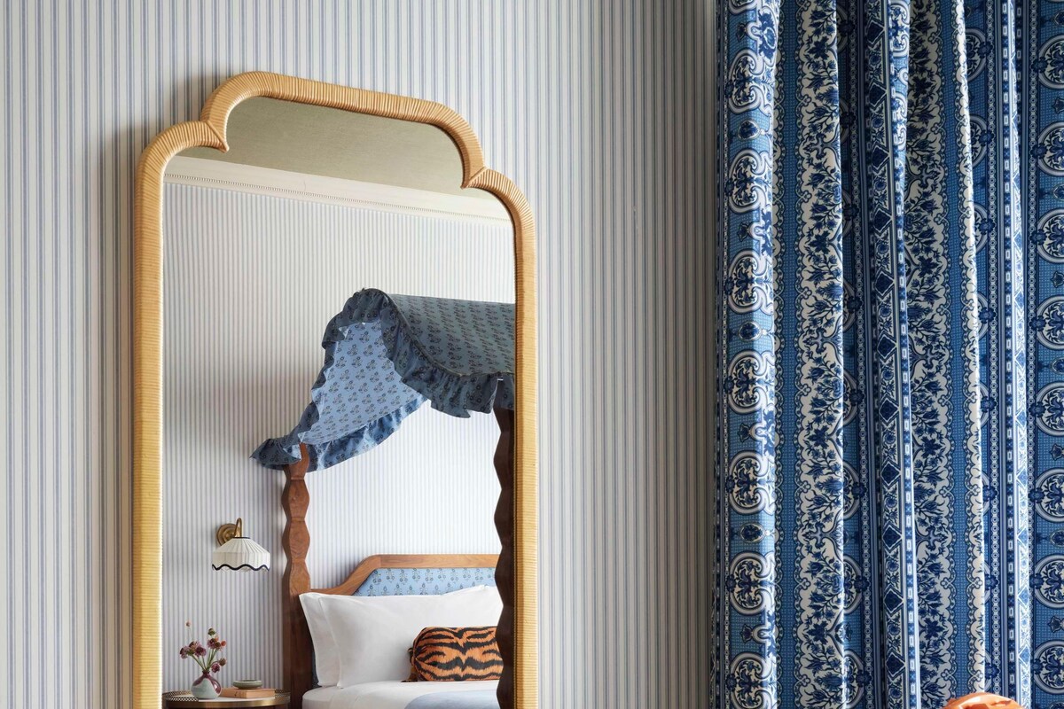 A room with a large mirror, a bed with a blue canopy, a wooden side table, and a patterned curtain.
