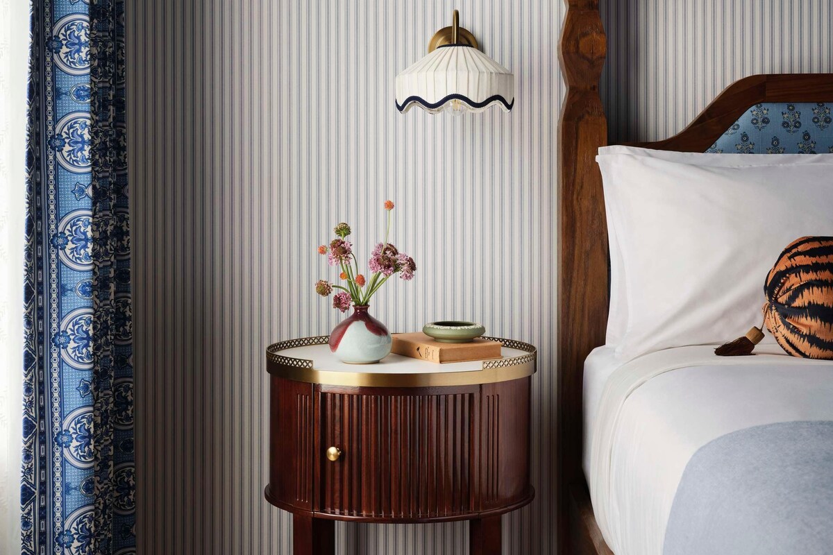 A bedroom with a wooden bed, a bedside table with a vase of flowers, and a wall-mounted lamp.