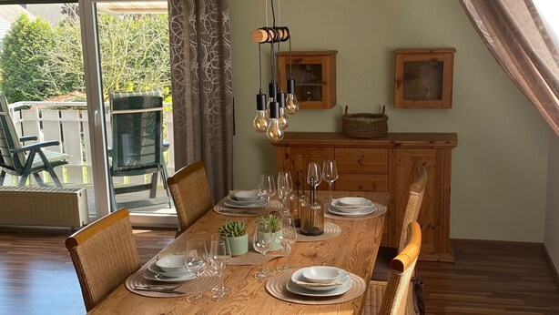 A dining area with a wooden table set for four, surrounded by wicker chairs, a wooden cabinet, and a hanging light fixture. - Ferienwohnung an der Schlösserachse . Ideal für Ausflüge ins Münsterland (Nordkirchen)