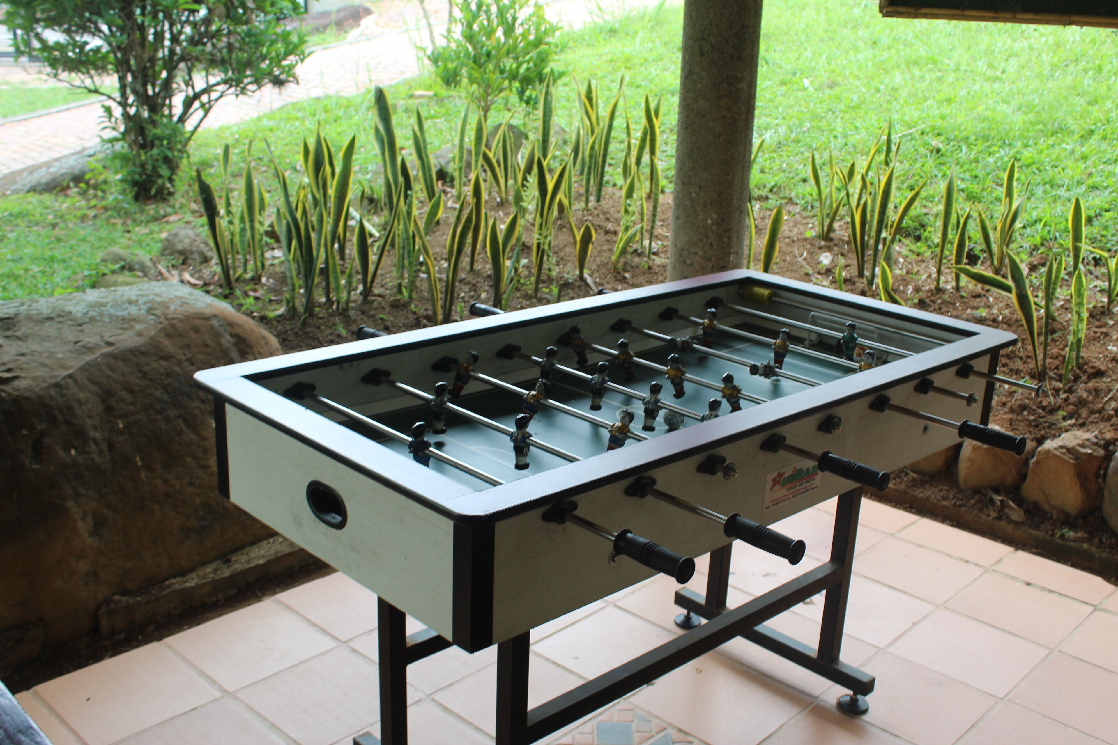 Una mesa de fútbol de mesa sobre un patio con baldosas, un área con césped y rocas al fondo.
