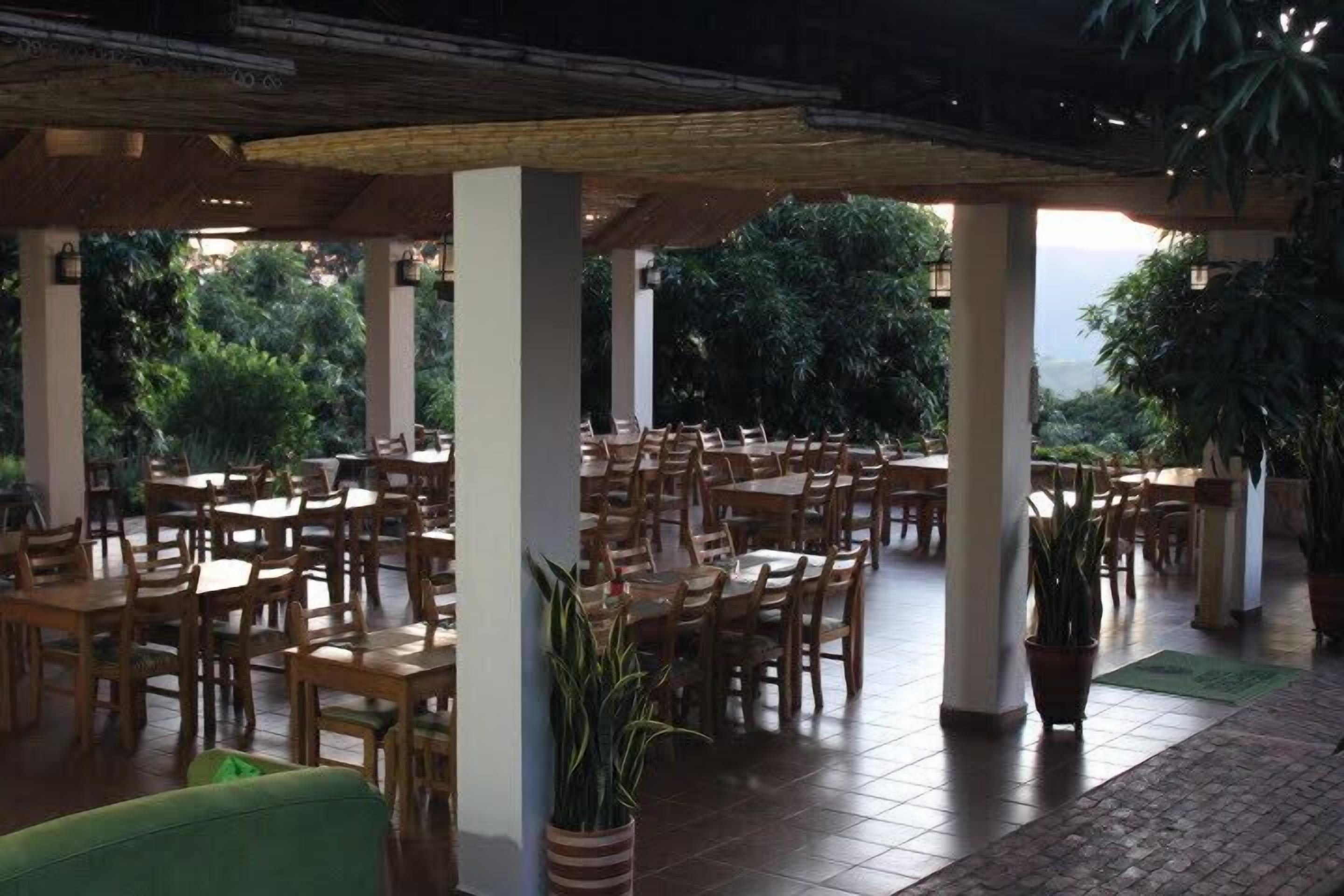 An outdoor dining area with wooden tables and chairs, potted plants, and a covered section with columns.