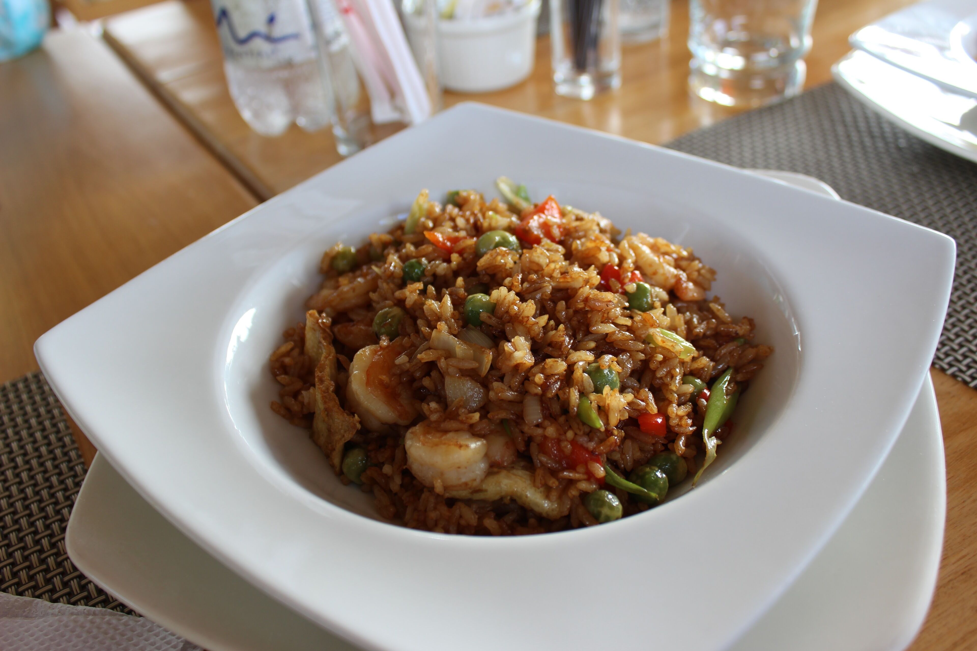 A white plate of fried rice with vegetables and shrimp on a wooden table.