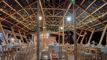 Un espace de restauration intérieur avec des tables et des chaises en bois, un plafond haut avec une structure en bambou et des guirlandes lumineuses.