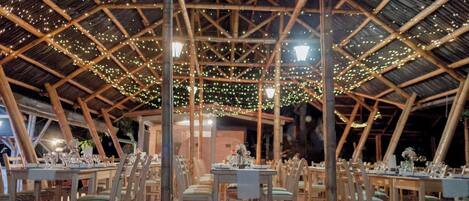 Un comedor interior con mesas y sillas de madera, un techo alto con estructura de bambú y luces colgantes.