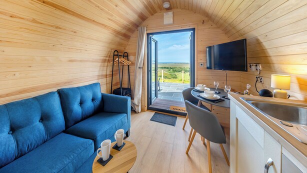 A cozy cabin interior with a blue sofa, a dining table with chairs, a TV mounted on the wall, and a view through the open door. - 1 bedroom accommodation in Talybont, near Borth (Talybont, near Borth)