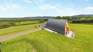 A modern, curved-roofed building with a paved entrance and a gravel path leading to it, set in a lush green field with distant hills under a clear sky. - 1 bedroom accommodation in Talybont, near Borth (Talybont, near Borth)
