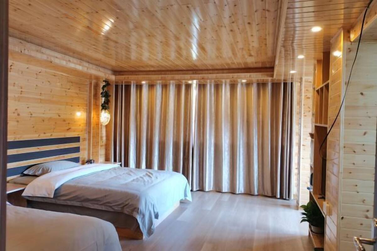 A wooden cabin-style room with two beds, a wooden ceiling, and a large window with curtains.