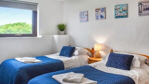 Hotellihuone, jossa on kaksi sänkyä, molemmissa siniset vuodevaatteet ja valkoiset tyynyt, pieni yöpöytä lampun kanssa ja ikkuna, josta avautuu näkymä vehreään maisemaan.