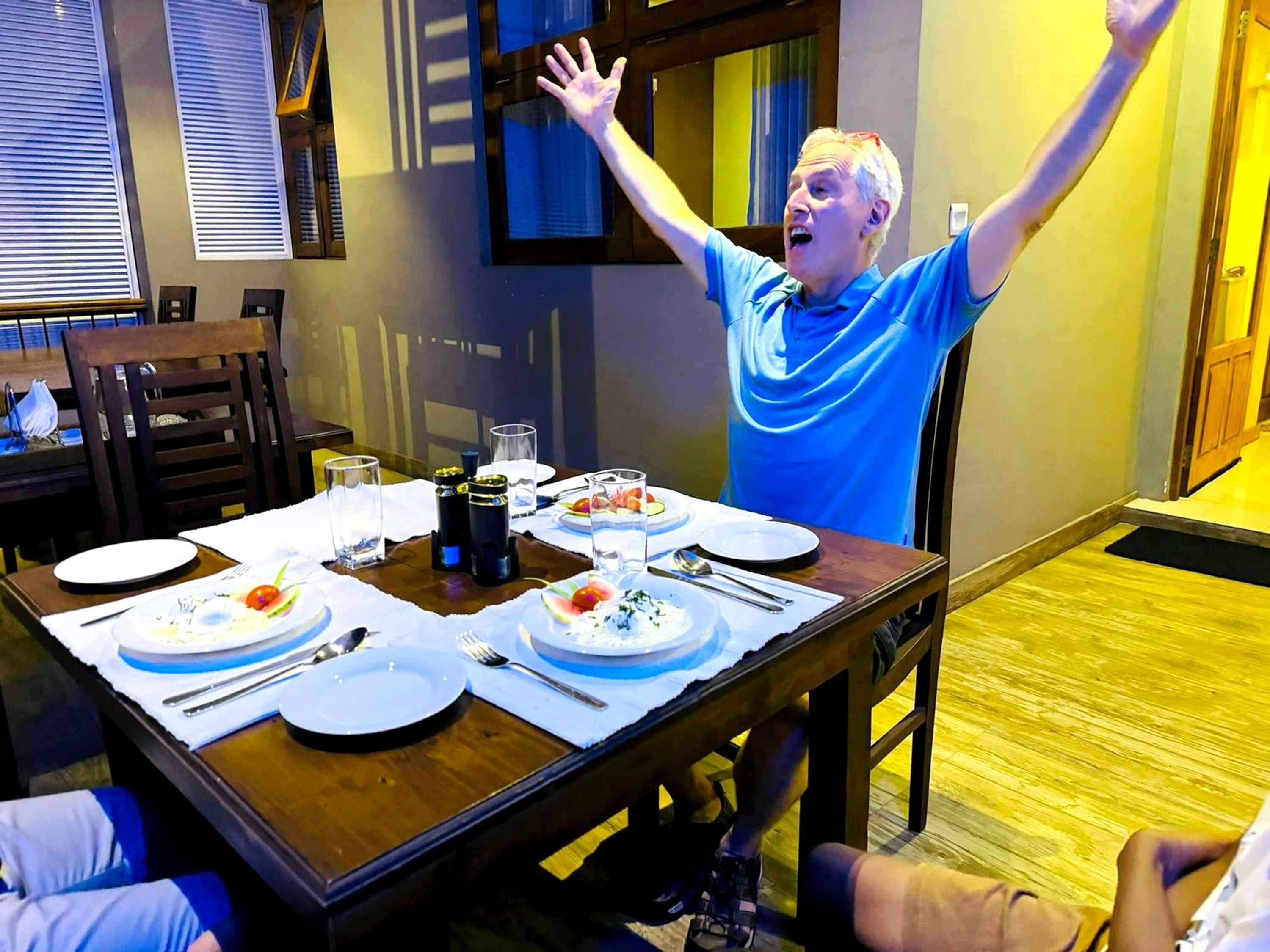 A dining table set with plates, glasses, and cutlery, with a person raising their arms in a room with wooden flooring and a window with blinds.