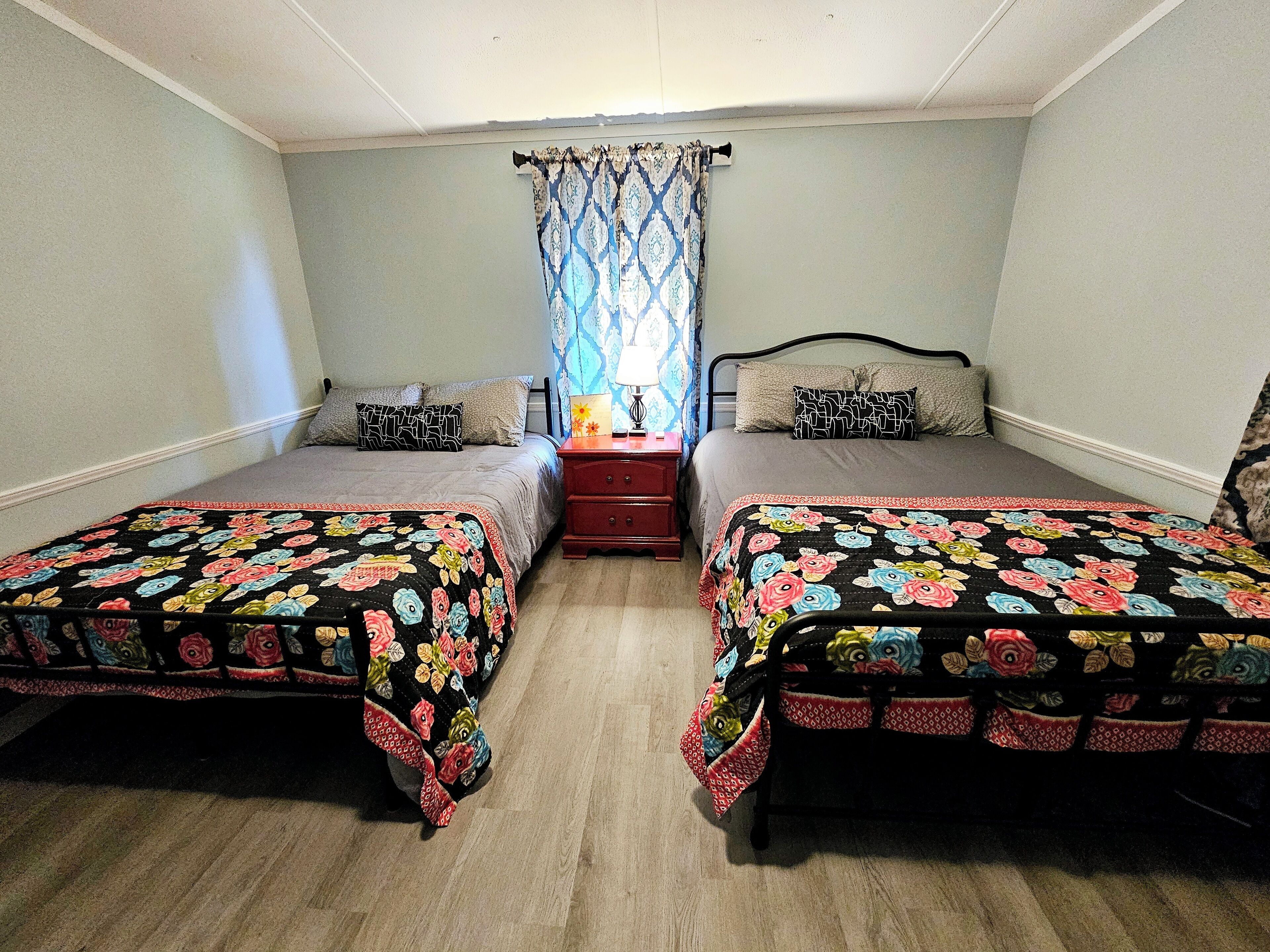 A bedroom with two beds, a nightstand, a lamp, and a window with curtains.