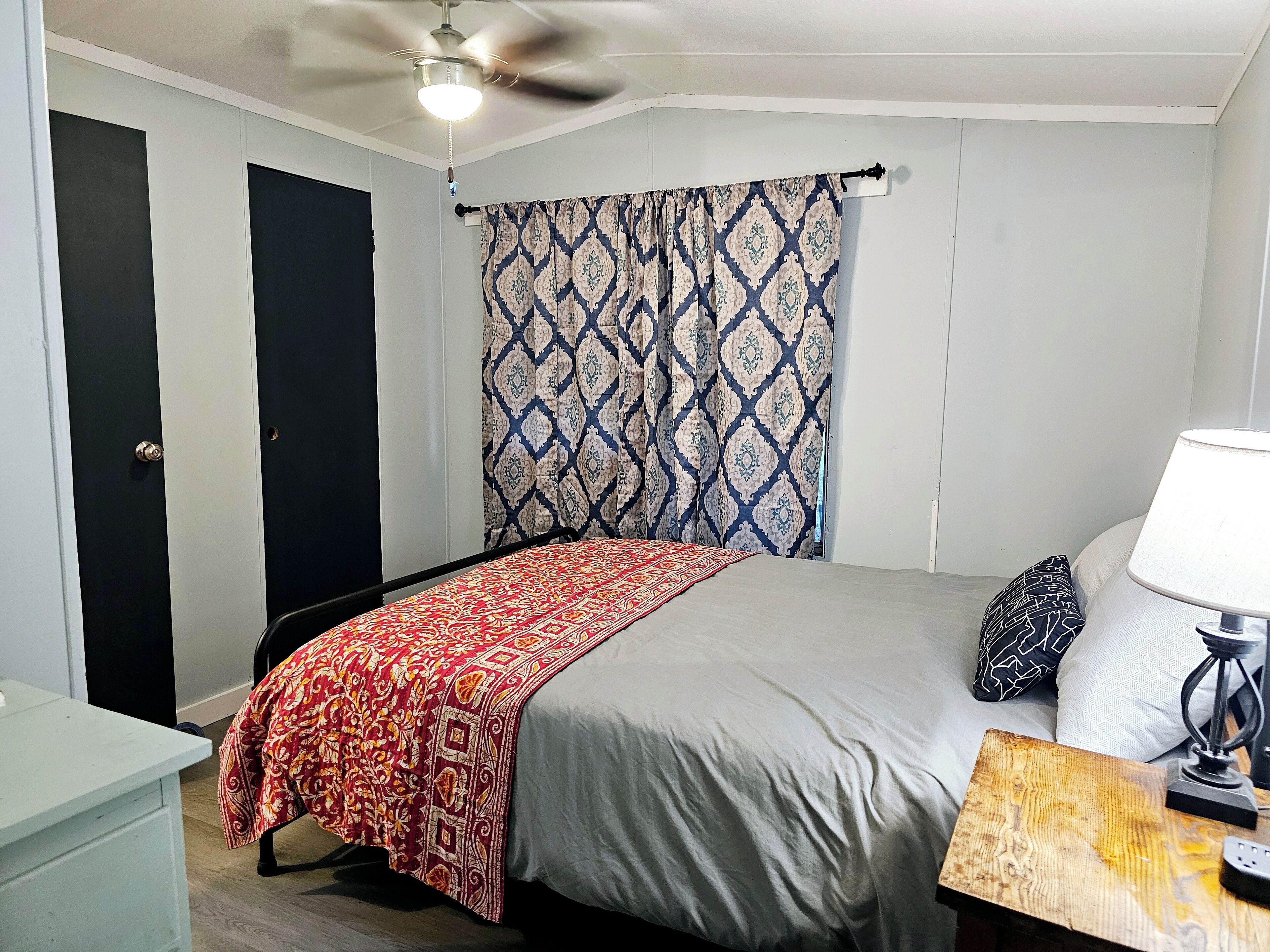 Ein Schlafzimmer mit einem Bett, einem Nachttisch mit Lampe, einem Deckenventilator und gemusterten Vorhängen.