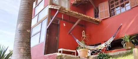 Un edificio rojo con una escalera de madera que lleva a una puerta, rodeado de follaje y una hamaca.