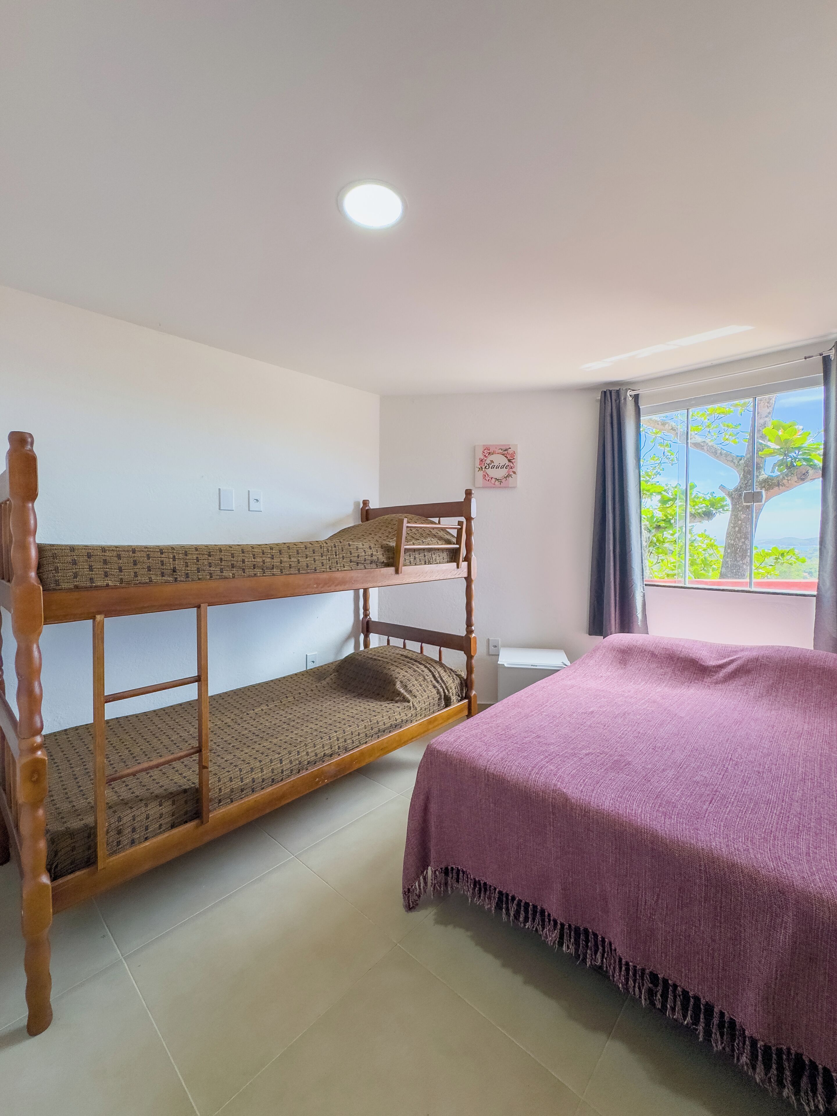 A room with a bunk bed and a single bed, a window with a view of trees, and a ceiling light.