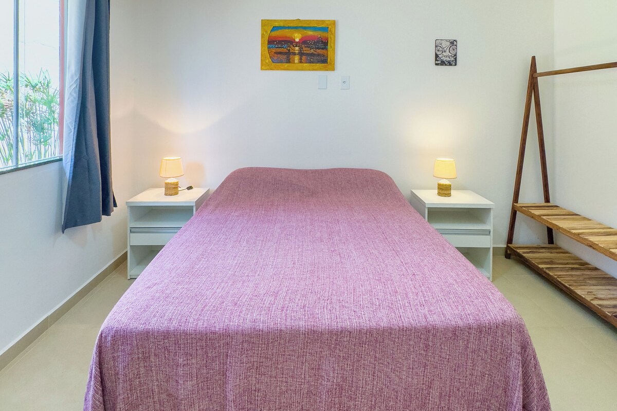 Une chambre à coucher avec un lit, deux tables de chevet avec des lampes, une fenêtre avec des rideaux, un tableau au mur et une étagère en bois.