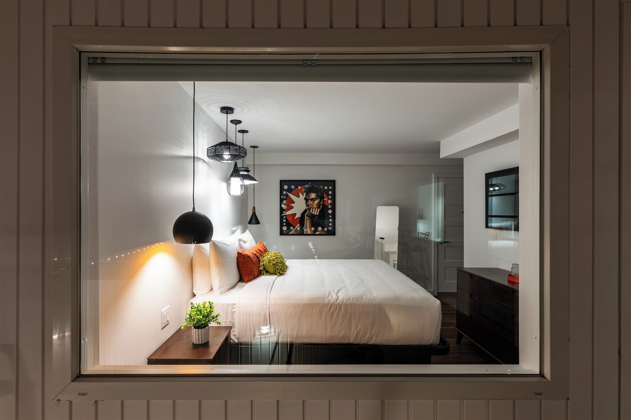 Quarto moderno com uma cama grande, abajures, uma mesinha com uma planta e um quadro na parede.