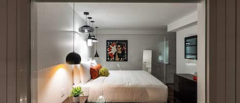 A modern bedroom with a large bed, bedside lamps, a small table with a plant, and a painting on the wall.