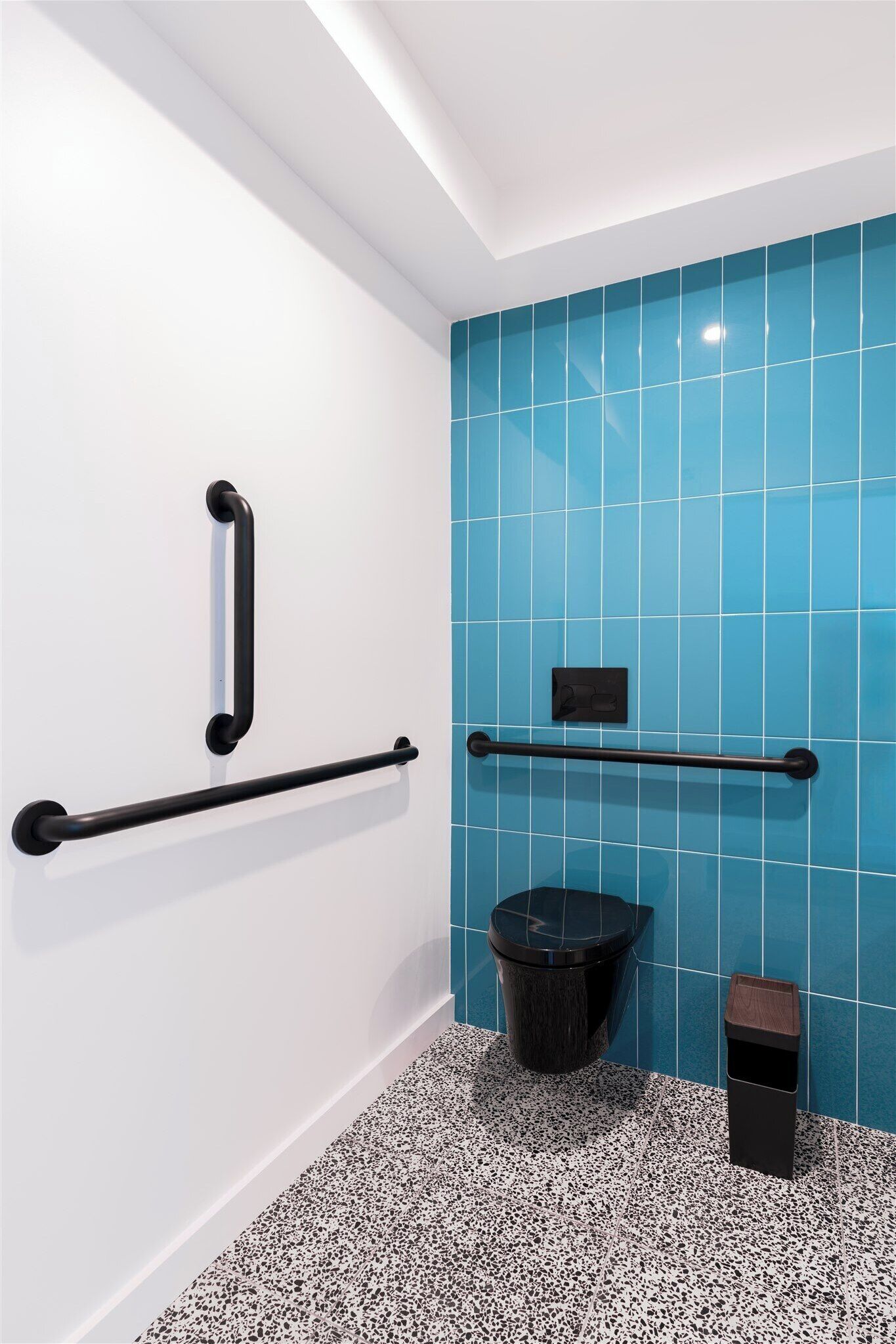 A bathroom with a white wall, blue tiled wall, black grab bars, a black trash bin, and a black toilet.