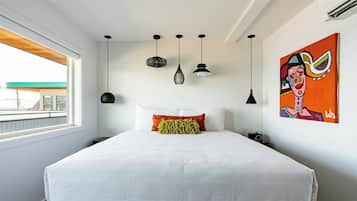 A modern bedroom with a large bed, multiple pendant lights, a vibrant painting, and a view of a building through a window.