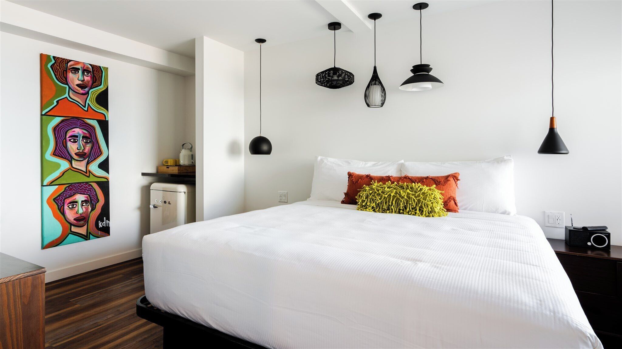 A modern bedroom with a large bed, white bedding, a colorful abstract painting, a small heater, and a black radio on the nightstand.